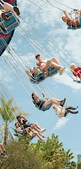 People riding a thrill swing ride in suspended seats with chains against a blue sky, looking happy and flying high.