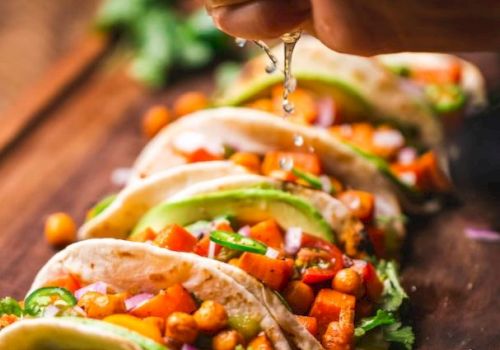 A person adds fresh toppings to tacos with veggies, cilantro, beans, and lime on a wooden table.