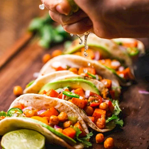 A person adds fresh toppings to tacos with veggies, cilantro, beans, and lime on a wooden table.