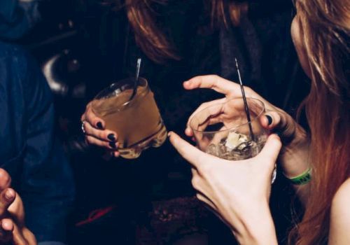 A party scene: young women hold cups with straws and cocktails, wearing dark nail polish and jewelry, laughing together.