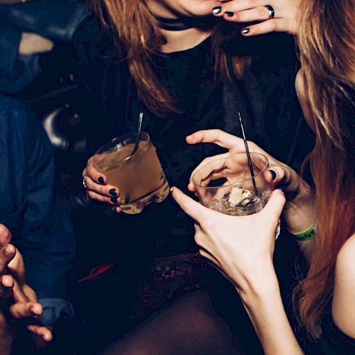 A party scene: young women hold cups with straws and cocktails, wearing dark nail polish and jewelry, laughing together.