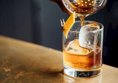 An amber cocktail being poured into a rocks glass over ice, garnished with a small pineapple spear and a slice.