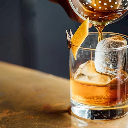 An amber cocktail being poured into a rocks glass over ice, garnished with a small pineapple spear and a slice.