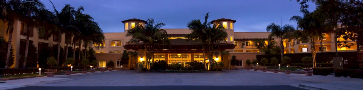 A resort or hotel courtyard at dusk, with tall palm trees and warmly lit buildings surrounding an open driveway.