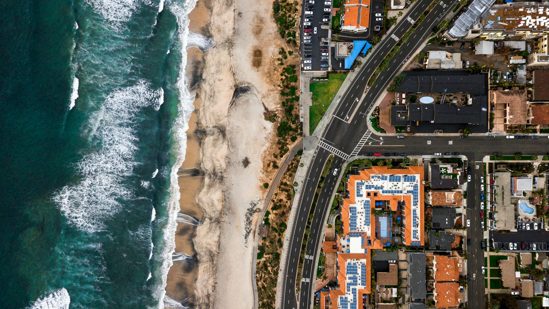 Coastal city scene: waves crash along a sandy beach next to a busy shoreline with roads, buildings, and parking lots on a cliffside.