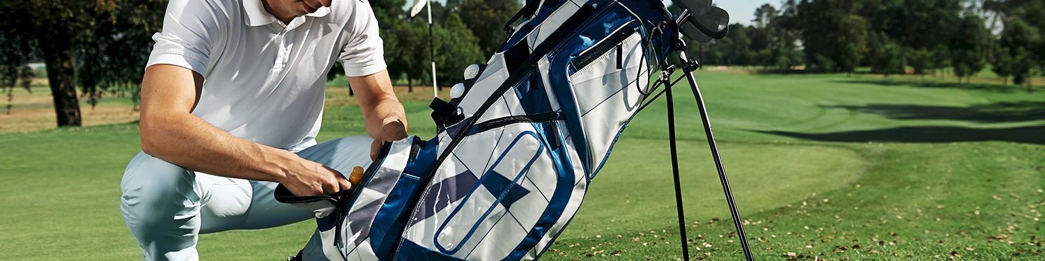 A man on a golf course is kneeling beside a large blue golf bag with clubs, adjusting or packing it, on a sunny day.