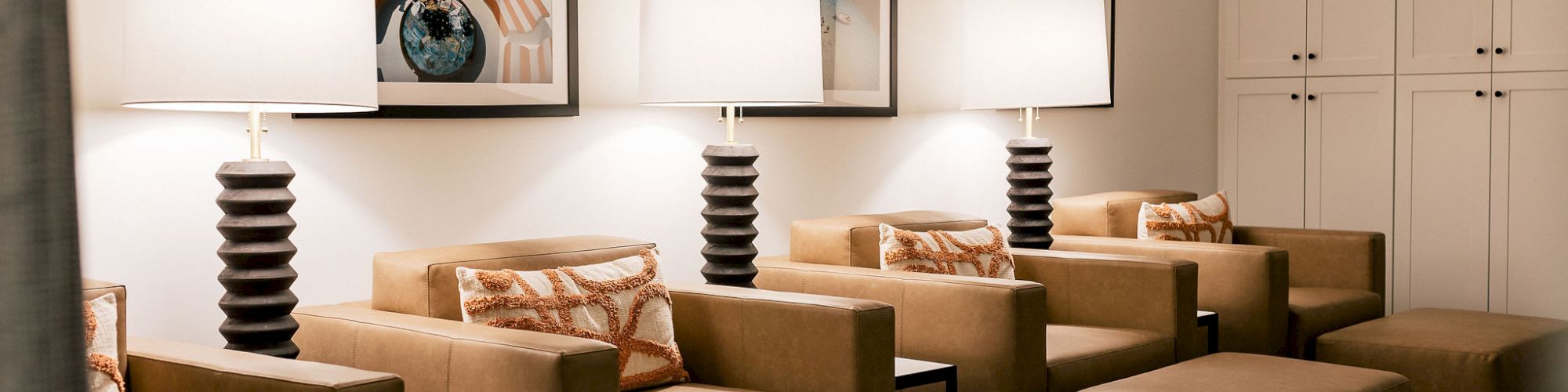 A stylish waiting lounge with six tan armchairs and matching ottomans, tall striped lamps, framed art, and built-in white cabinets in a calm setting.
