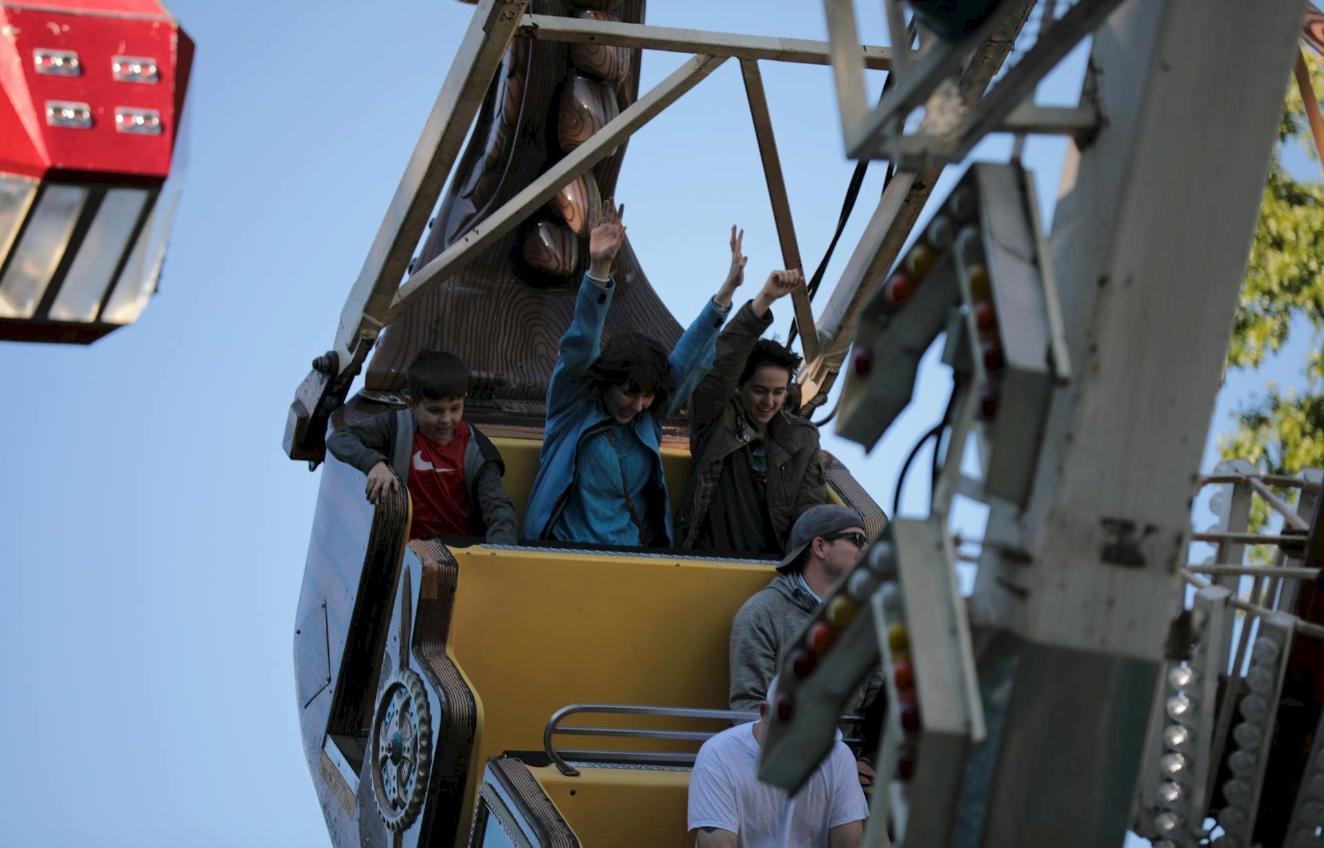 A group of people rides a Ferris wheel cart, waving and smiling as they ascend, with a red cabin nearby against a clear blue sky.
