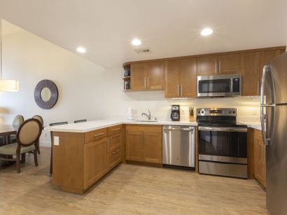 A modern kitchen with wooden cabinets, stainless steel appliances, a center island, and a small dining area to the left, bright and sunny.
