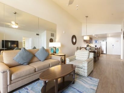 Bright open-plan living room with a beige sofa, coffee table, blue rug, and a view into the kitchen.