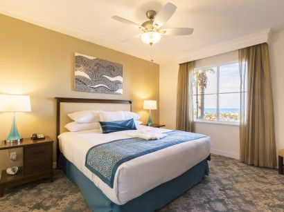 A neatly made hotel room with a king bed, teal accents, two nightstands, lamps, artwork, and a window overlooking a sunny view.