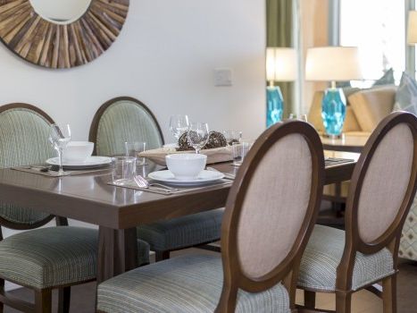 A stylish dining room with a wooden table set for a meal, upholstered chairs, polished place settings, and a decorative wall mirror in the background.