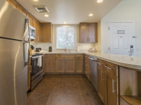 A modern U-shaped kitchen with wooden cabinets, stainless steel fridge and stove, light countertops, and a window over the sink, door to the right.