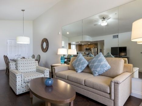 A modern living room with a beige sofa, blue patterned cushions, a dark wood coffee table, armchairs, mirrored wall, and soft lighting.