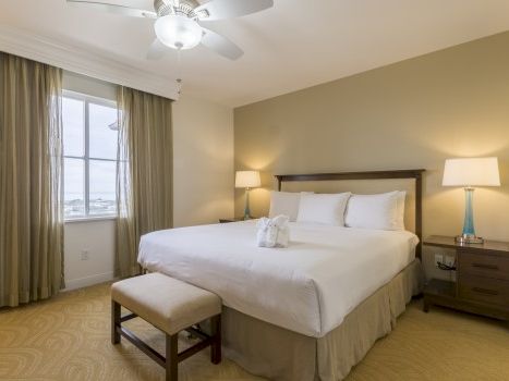 A neatly made hotel room with a king bed, two nightstands, lamps, a small bench, beige walls, and a window with heavy curtains.