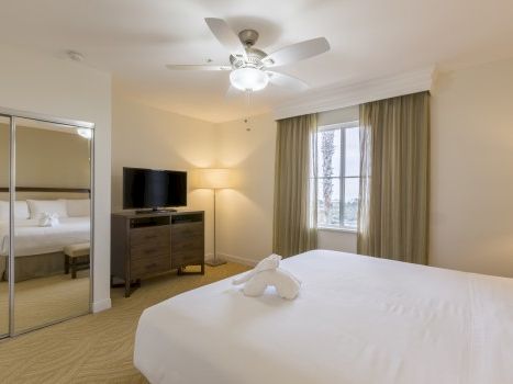 A bright modern bedroom with a king bed, ceiling fan, TV stand, curtains, window, and a towel folded on the bed.