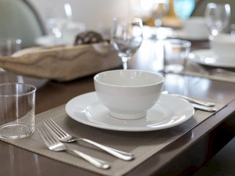 A dining table set for a meal with a white bowl on a plate, glassware, cutlery, napkin, and a soft, blurred background.