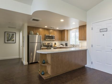 Cozy kitchen with stainless fridge, wooden cabinets, island counter, and a hallway leading to a doorway, all under soft overhead lighting.