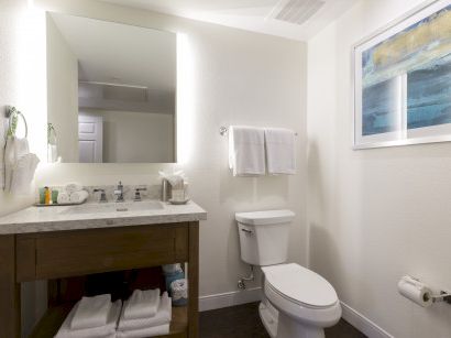 A modern bathroom with a wooden vanity, sink, mirror with backlighting, toilet, towel rack, artwork, and a few toiletries near the sink.