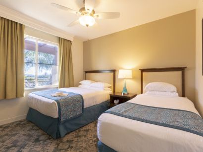 Two twin beds in a bright hotel room with a nightstand, lamp, ceiling fan, and a window with tan curtains. The d&eacute;cor is beige and teal, neat and cozy.