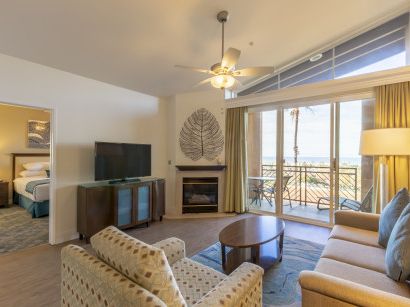 A bright hotel living room with a fireplace, sofa and chairs, a TV on a cabinet, large windows with a balcony, and a view outside.