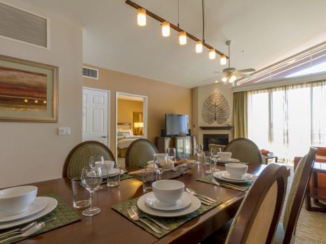 A stylish dining room with a long table set for eight, place settings, glassware, and modern lighting; living area and large window on the right.