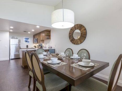 A modern dining area with a dark wood table set for four, adjacent to a bright open kitchen; a round wall mirror and a large pendant light above.