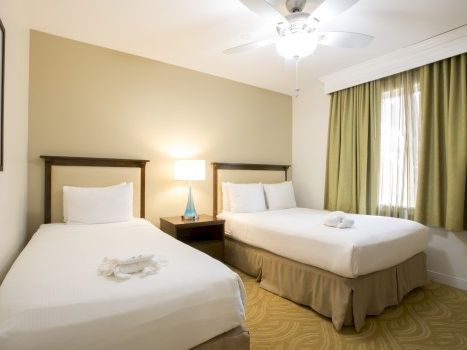 A hotel room with two queen beds, a nightstand, lamp, beige walls, and green curtains, neatly made and inviting.
