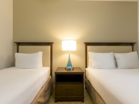 Two twin beds with white linens flanking a shared nightstand and lamp, against a light olive wall.