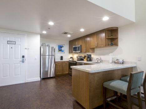 A modern kitchen with a stainless fridge, wooden cabinets, a white island, bar seating, and pendant lights in a bright, open space.