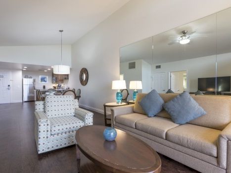 A bright, modern living room with a beige sofa and patterned armchairs, a round coffee table, and a large wall mirror reflecting a tidy, open-concept space.