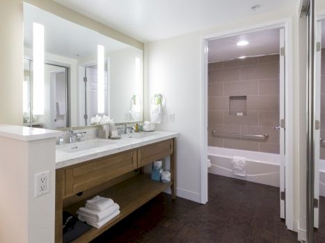 A modern hotel bathroom with a double sink vanity, mirrors and lighting, open shelving under the sink, and a separate shower and bathtub area in a clean, airy space.