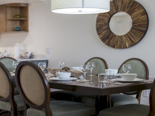 A modern dining room with a dark wooden table set for a meal, six upholstered chairs, glassware and bowls, a round sunburst wall mirror, and a kitchen area in the background.