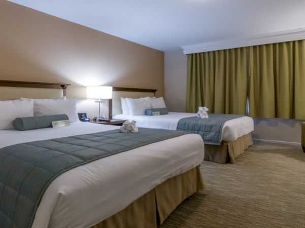 Two double beds in a hotel room with teal bed runners, a bedside lamp, a sitting chair, and gold curtains, creating a cozy, neutral-toned space.