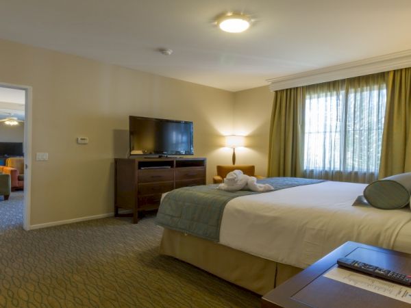 A hotel suite with two queen beds, a dresser with TV, and a seating area visible through an open doorway, in soft beige tones.