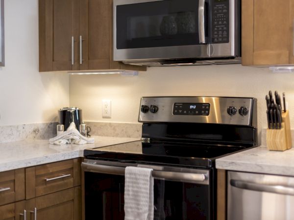 A tidy kitchen with wooden cabinets, stainless steel stove/oven, microwave, marble countertop, small appliances, and a knife block on the counter.