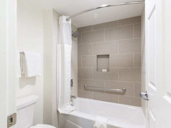 A modern bathroom with a tub and shower combo, gray tiled wall, curved shower rod, towel rack, and white fixtures, clean and fresh.