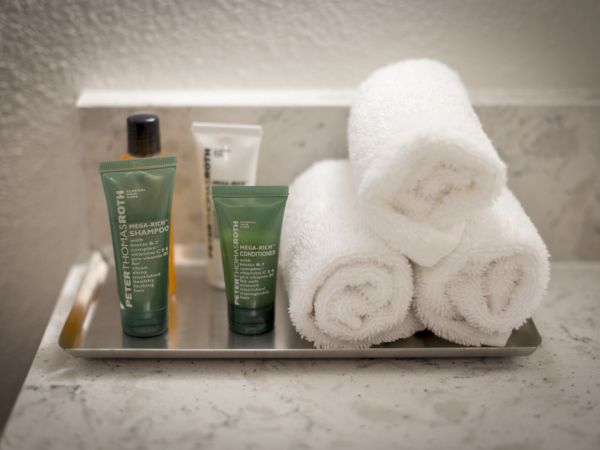 A metal tray holds three green skincare tubes and a small bottle, plus three neatly rolled white towels on a marble counter.