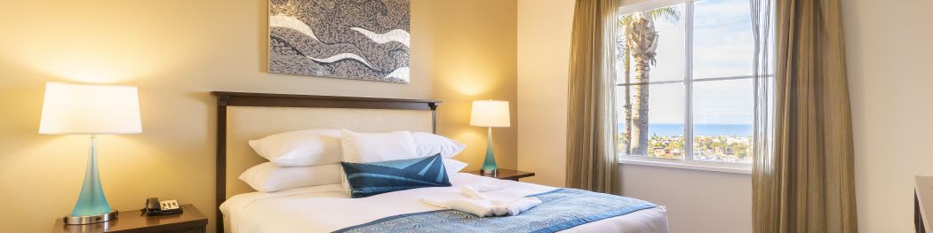 A hotel room with a king bed, teal accents, two bedside tables with lamps, wall art, a ceiling fan, and a window view to the outside.
