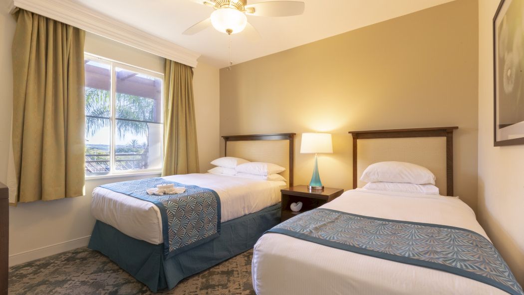 A hotel room with two double beds, beige walls, a nightstand with a lamp, a ceiling fan, and a window with green curtains.