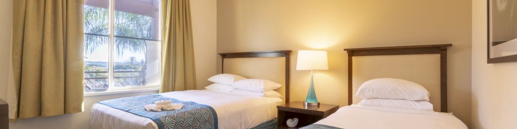 A hotel room with two double beds, beige walls, a nightstand with a lamp, a ceiling fan, and a window with green curtains.