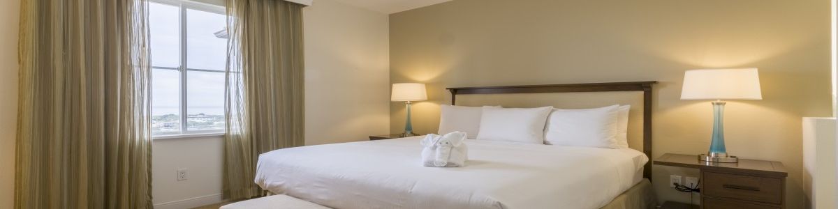 A plush hotel room with a large king bed, nightstands, two lamps, a bench at foot, beige walls, and a window with green curtains, bright and tidy.