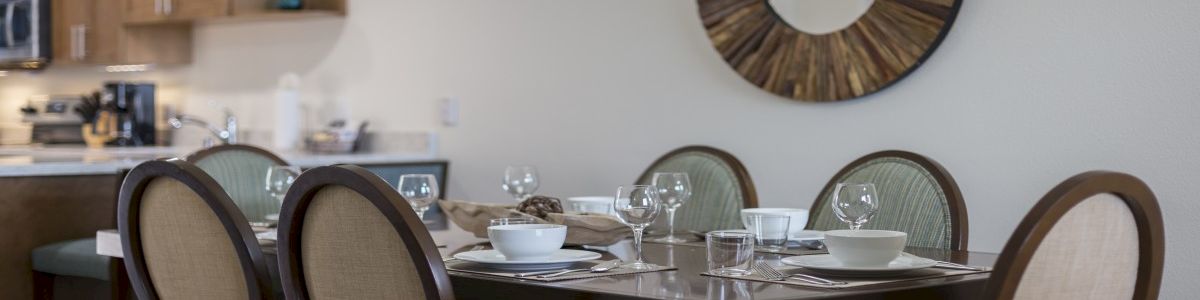 A modern dining room with a dark wood table set for six, oval-backed chairs, glassware and bowls, a circular sunburst mirror, and a kitchen area in the background.