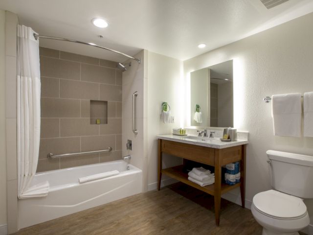 A modern bathroom featuring a bathtub with a shower curtain, glass door, tiled surround, a wooden vanity with sink, mirror, towels, and a toilet.