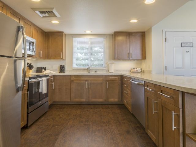 A compact kitchen with wooden cabinets, a stove, sink, tile backsplash, and a windowed doorway at the far end, all in a warm, neutral tone.