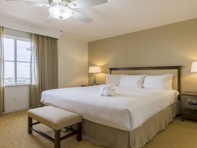 A bright hotel room with a large bed, side tables, a chair, desk, and a window with curtains, plus warm lighting.
