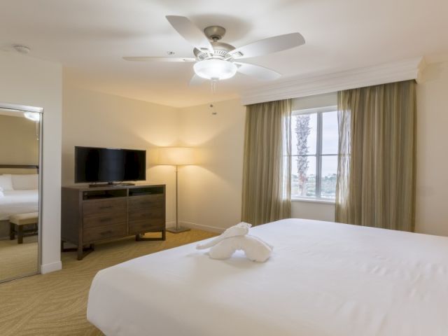A hotel room with a bed in the foreground, a desk and chair, a flat-screen TV, and large windows with curtains, soft lighting.