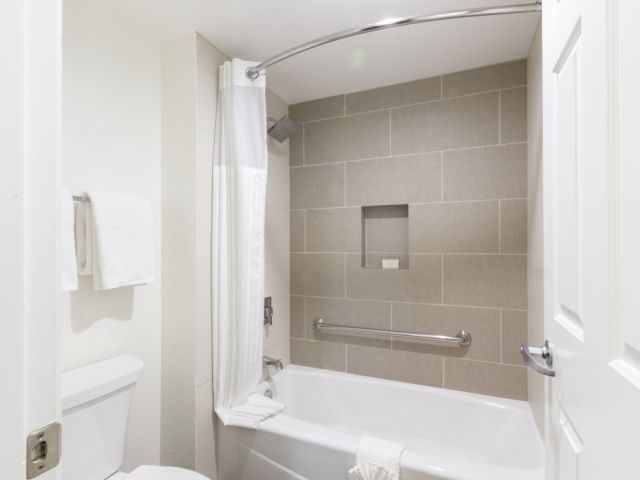 A modern bathroom with a white toilet, tiled gray shower walls, a curved shower rod, a built-in niche, and a safety grab bar by the tub, neat.
