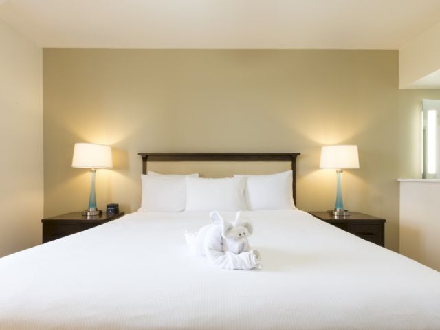A tidy hotel bedroom with a large white bed, two matching nightstands, two lamps, a towel animal centerpiece, and a soft beige accent wall.