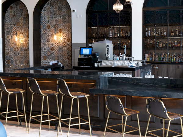 A modern coffee bar with espresso machine, glassware, stools, and ornate arched tile panels behind it.